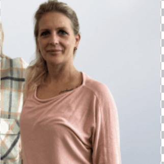 Woman with blonde hair wearing a pink long-sleeve top, standing indoors and facing the camera (partial person in plaid shirt visible to the left).